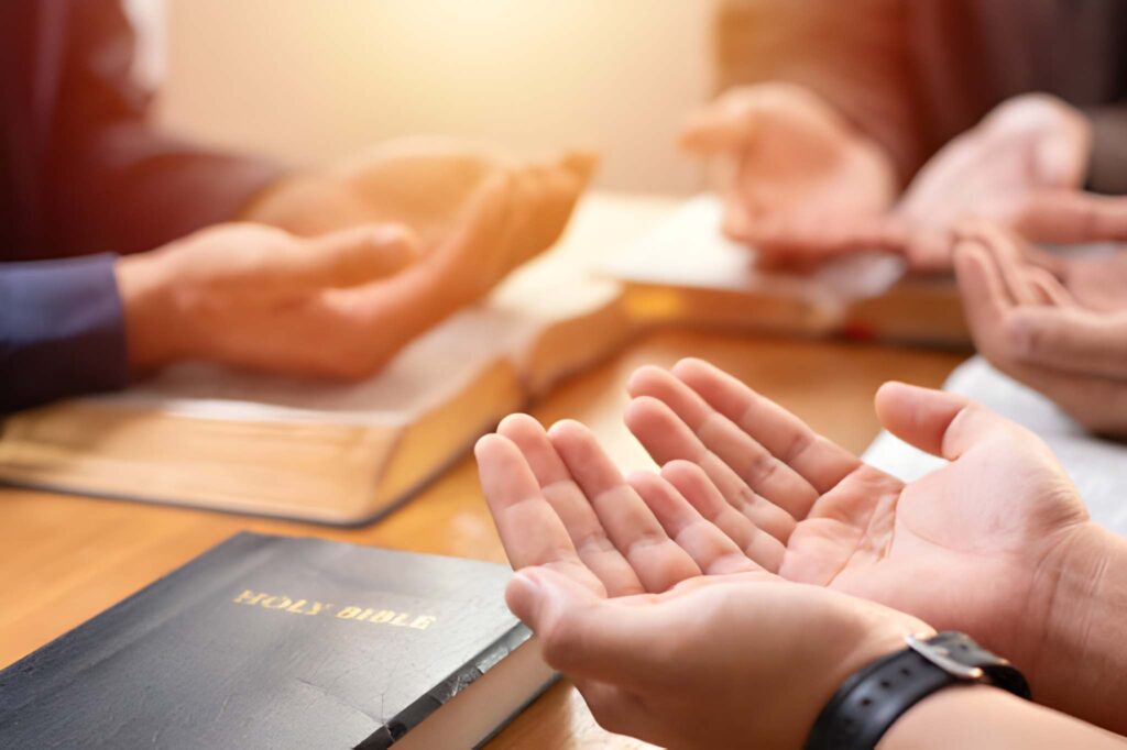 A group of people are gathered around a table with their hands outstretched in thanksgiving, with Bibles on the table